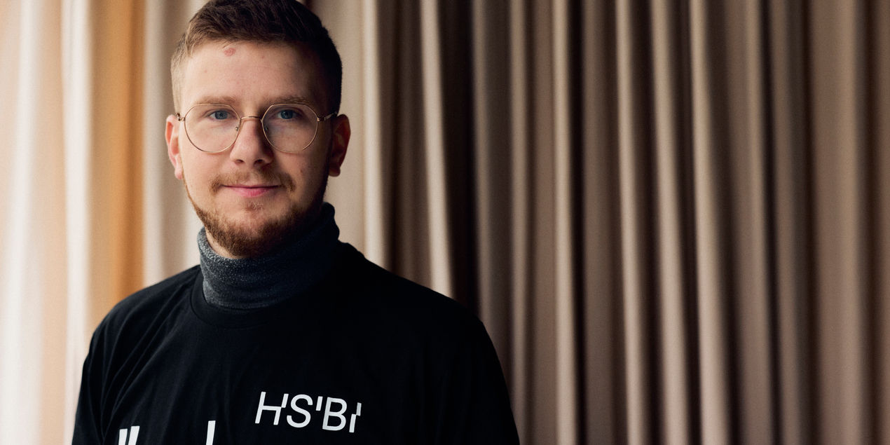 Porträtbild von Samuel Wiebe Er steht in einem schwarzen T Shirt mit HSBI Aufdruck vor einem rosafarbenen Vorhang im Berufskolleg Senne Sein markantes Gesicht wird durch den Backenbart und seine Brille mit schamalem Rand geprägt