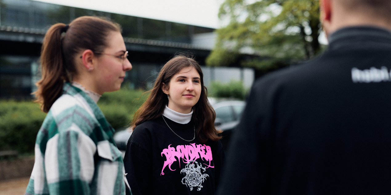 Bild von maya Chrobok die Schülerin am Berufskolleg Senne ist Sie steht in einem schwarzen T Shirt auf dem Schulhof neben ihr steht eine Mitschülerin ihr gegenüber mit dem Rücken zur Kamera erkennt man Samuel Wiebe